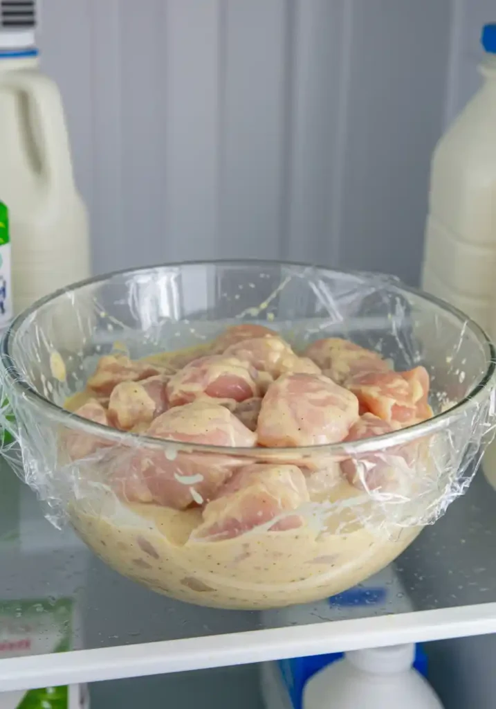 Raw chicken pieces marinating in a bowl of seasoned liquid, covered with plastic wrap in a refrigerator